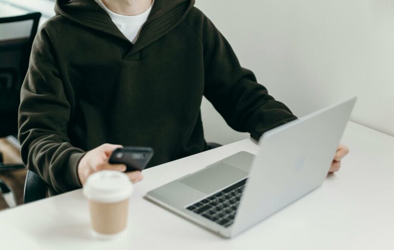 Casually dressed man working from home using a laptop and smartphone, with a coffee cup nearby.