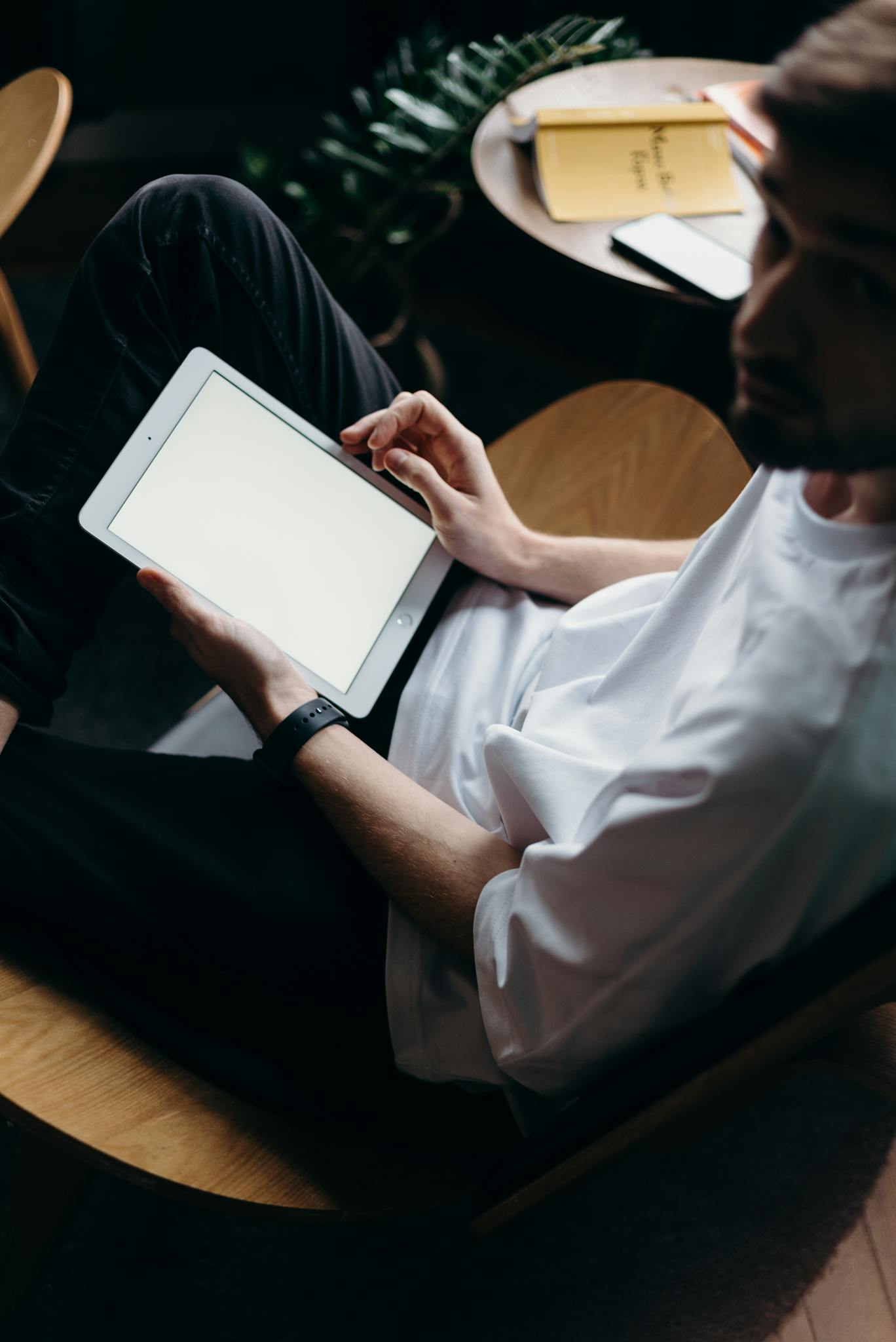 Man wearing white T-shirt using a tablet while relaxing in a cozy home setting.