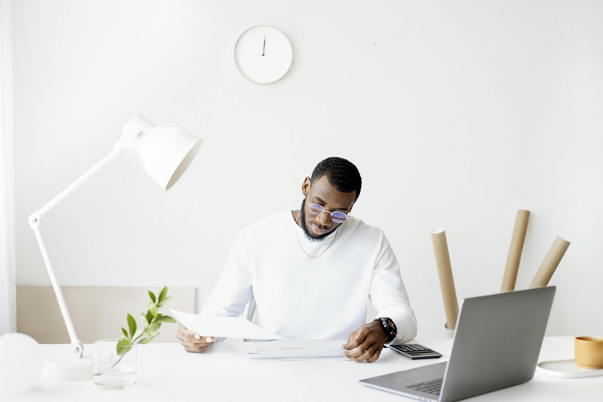 A professional adult working intently at a minimalist desk with a laptop, papers, and a lamp.