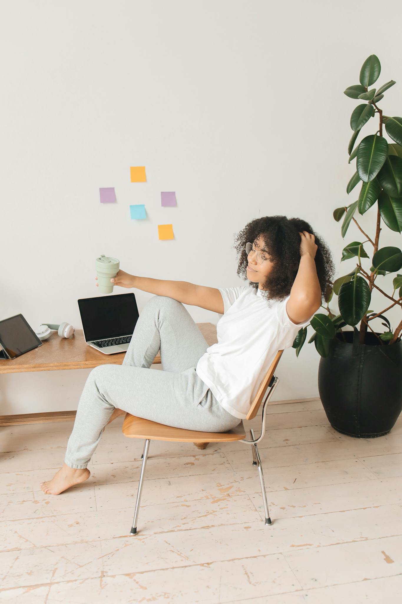 A woman in casual attire relaxing with a laptop and drink in a home office with indoor plants.