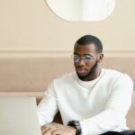 Black male professional focused on laptop in modern office setting, wearing glasses and casual attire.
